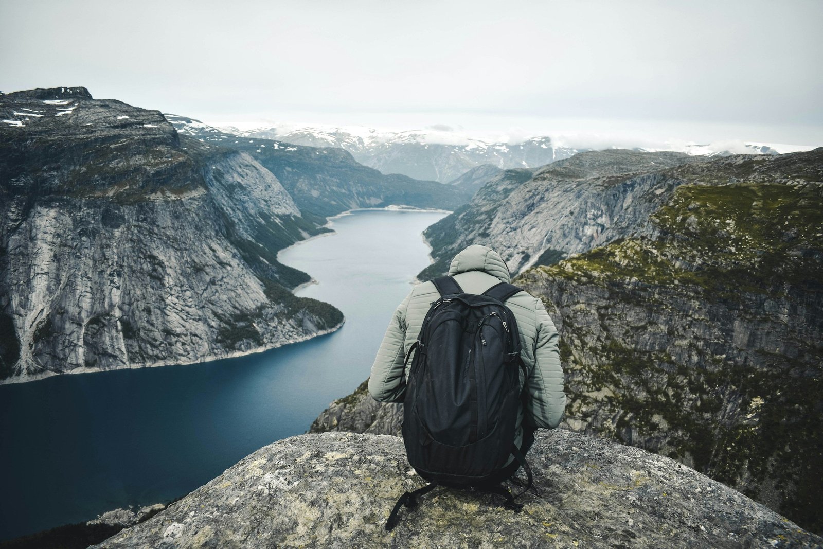 A hiker with a black backpack sits on a rock ledge above a fjord in Norway.