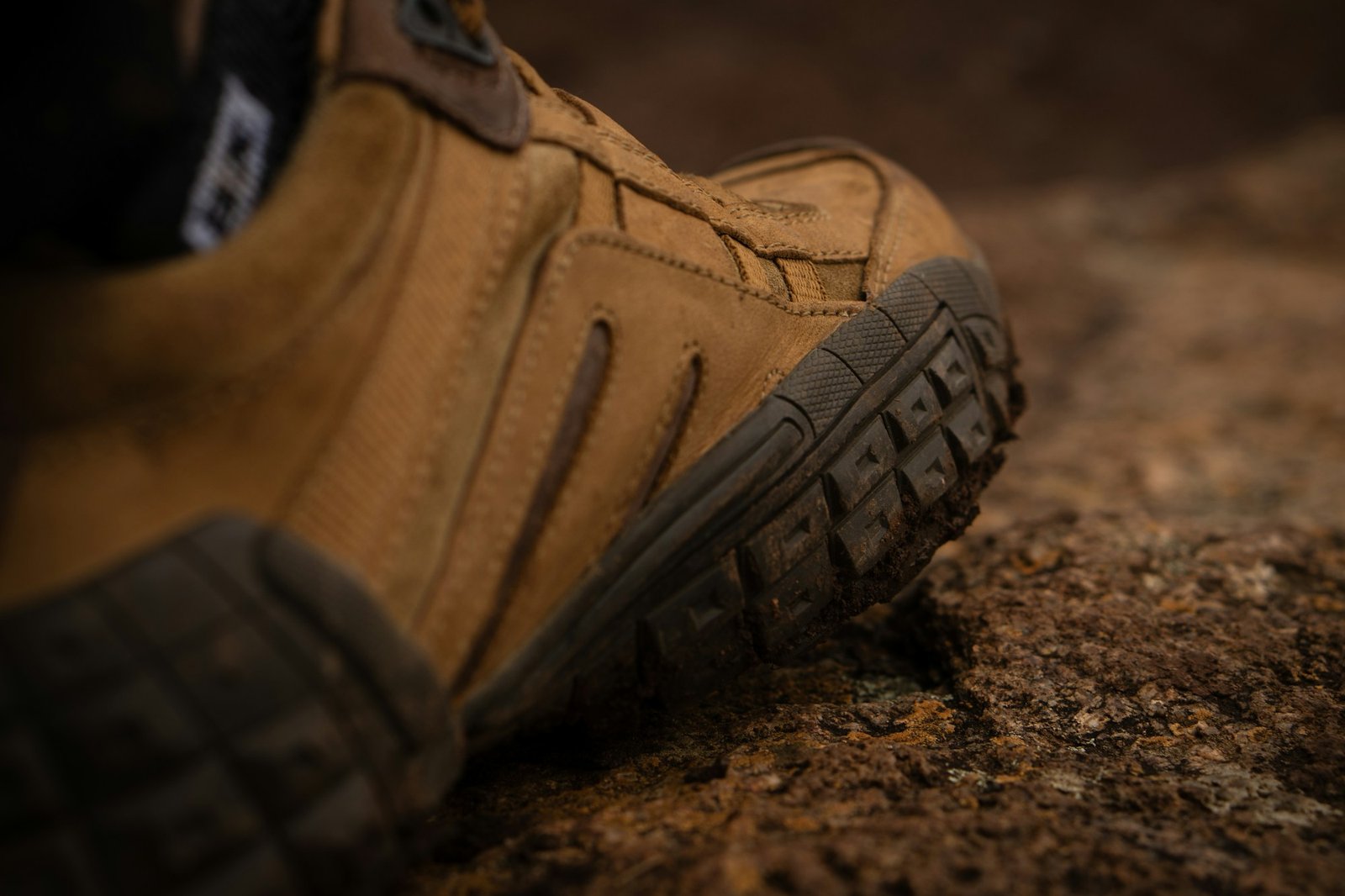 Close-up of a tan boot with a deep lugged rubber outsole on dirt.